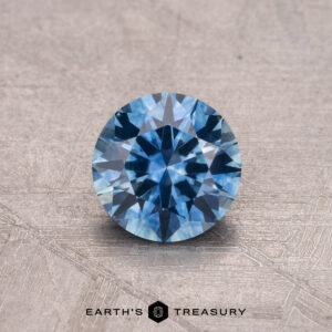 A faceted 0.89-carat blue Montana Sapphire is showcased on a textured surface, accompanied by the "Earth's Treasury" logo text.
