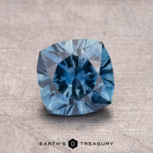 A faceted cushion-cut blue 1.27-carat Montana Sapphire (heated) on a textured surface with the text "Earth's Treasury" below.