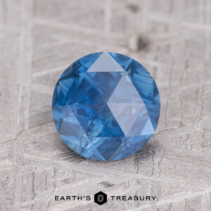 A round MS7403 gemstone in blue, positioned on a textured surface.