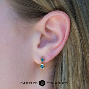 Close-up of a person's ear wearing Dewdrop earrings featuring double blue gemstones set in gold, with visible light brown hair. The EARTH'S TREASURY logo appears at the bottom.