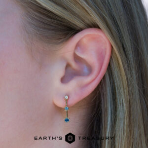 Close-up of a person's ear wearing the elegant dusk earring with two colored stones set in gold. The "EARTH'S TREASURY" logo appears at the bottom of the image.