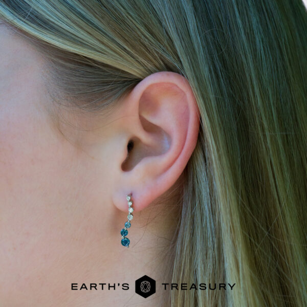 journey Close-up of a woman's ear with straight, light hair, wearing the Journey earrings—curved and set with multiple blue gemstones. The "EARTH'S TREASURY" logo is displayed at the bottom.