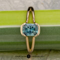The "Madi" ring in 18K yellow gold with a 1.41-carat Montana sapphire is displayed vertically on a green and silver textured surface, with "Earth's Treasury" written at the bottom.