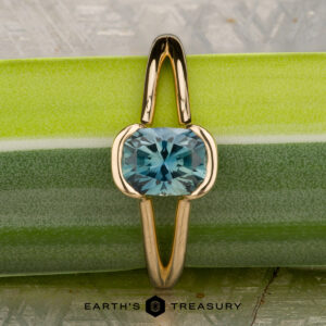 The "Madi" ring in 18K yellow gold with a 1.41-carat Montana sapphire is displayed vertically on a green and silver textured surface, with "Earth's Treasury" written at the bottom.