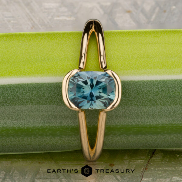 The "Madi" ring in 18K yellow gold with a 1.41-carat Montana sapphire is displayed vertically on a green and silver textured surface, with "Earth's Treasury" written at the bottom.
