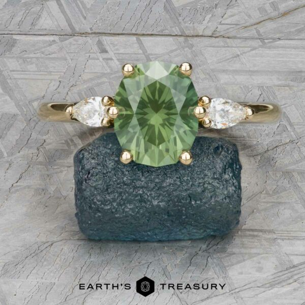 The "Nymph" ring in 18k yellow gold with 2.74-carat Australian sapphire The "Nymph" ring in 18k yellow gold, featuring a 2.74-carat Australian sapphire and two pear-shaped clear stones, is displayed on a rough blue mineral with the "Earth's Treasury" logo below.