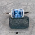 A "Lina" ring in 14k white gold with a 2.34-carat Montana sapphire is displayed on a textured blue stone, with "EARTH'S TREASURY" text below.