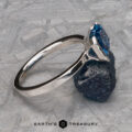 The "Lina" ring in 14k white gold with a 2.34-carat Montana Sapphire rests on a rough dark mineral surface. The light gray background features intersecting lines, and "EARTH'S TREASURY" appears at the bottom.