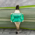 The "Hera" ring in 18k yellow gold features a 2.88-carat rectangular emerald center stone and two trapezoid-cut side diamonds, displayed on a green striped surface.