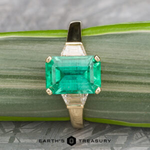 The "Hera" ring in 18k yellow gold features a 2.88-carat rectangular emerald center stone and two trapezoid-cut side diamonds, displayed on a green striped surface.