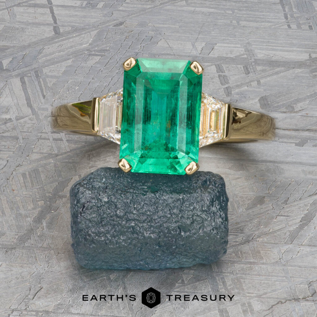 The "Hera" ring in 18k yellow gold, featuring a 2.88-carat emerald and two side stones, is displayed on raw dark green mineral with the "Earth's Treasury" logo below.