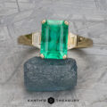 The "Hera" ring in 18k yellow gold, featuring a 2.88-carat emerald and two side stones, is displayed on raw dark green mineral with the "Earth's Treasury" logo below.