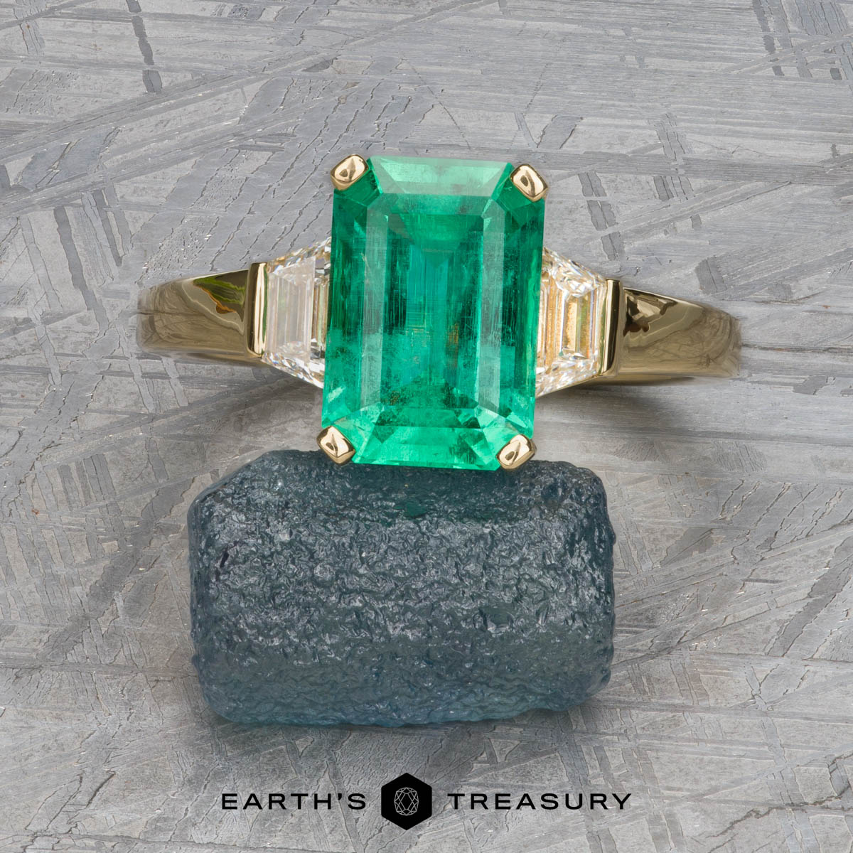 The "Hera" ring in 18k yellow gold, featuring a 2.88-carat emerald cut gemstone with side stones, showcased on a rough stone block with "Earth's Treasury" beneath.