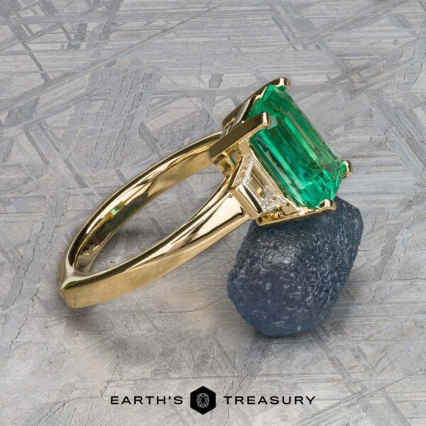 The "Hera" ring in 18k yellow gold with 2.88-carat emerald The "Hera" ring in 18k yellow gold features a 2.88-carat emerald with small diamonds on the sides, displayed on a rough stone. The Earth’s Treasury logo is positioned at the bottom center.
