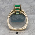 The "Hera" ring in 18k yellow gold with a 2.88-carat emerald is displayed upright, highlighting its side setting against a geometric background with the text "EARTH'S TREASURY" at the bottom.