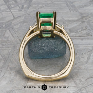 The "Hera" ring in 18k yellow gold with a 2.88-carat emerald is displayed upright, highlighting its side setting against a geometric background with the text "EARTH'S TREASURY" at the bottom.