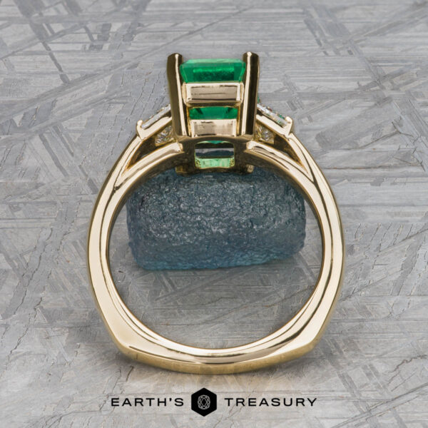 The "Hera" ring in 18k yellow gold with 2.88-carat emerald The "Hera" ring in 18k yellow gold with a 2.88-carat emerald is displayed upright, highlighting its side setting against a geometric background with the text "EARTH'S TREASURY" at the bottom.