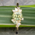 The "Liana" ring in 18k yellow gold features a 1.50-carat round Australian sapphire at its center, accented by marquise and round white stones, shown resting on a green leaf.