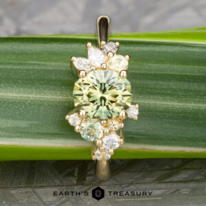 The "Liana" ring in 18k yellow gold features a 1.50-carat round Australian sapphire at its center, accented by marquise and round white stones, shown resting on a green leaf.