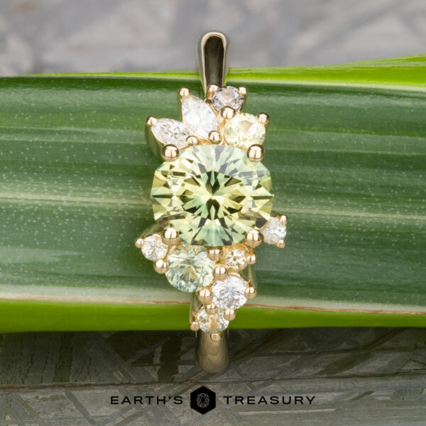 The "Liana" ring in 18k yellow gold features a 1.50-carat round Australian sapphire at its center, accented by marquise and round white stones, shown resting on a green leaf.