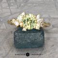 The "Liana" ring in 18k yellow gold features a 1.50-carat Australian sapphire accented by smaller clear and green stones, shown on a rough green mineral with the "Earth's Treasury" logo below.