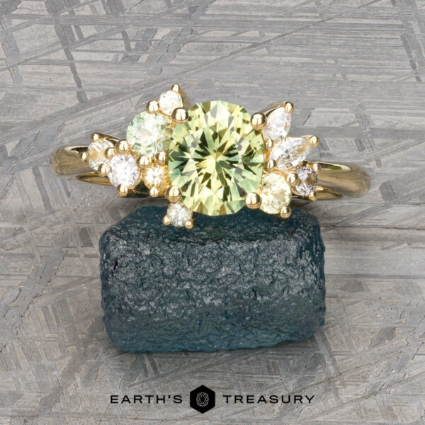 The "Liana" ring in 18k yellow gold with 1.50-carat Australian sapphire The "Liana" ring in 18k yellow gold features a 1.50-carat Australian sapphire accented by smaller clear and green stones, shown on a rough green mineral with the "Earth's Treasury" logo below.