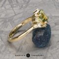 The "Liana" ring in 18k yellow gold, featuring a 1.50-carat Australian sapphire, rests against a rough, dark blue mineral on a patterned gray surface.