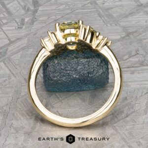 The "Liana" ring in 18k yellow gold with a 1.50-carat Australian sapphire stands upright on a rough blue stone, set against a geometric patterned background with "EARTH'S TREASURY" at the bottom.
