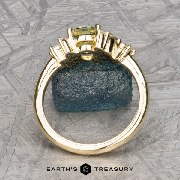 The "Liana" ring in 18k yellow gold with 1.50-carat Australian s The "Liana" ring in 18k yellow gold with a 1.50-carat Australian sapphire stands upright on a rough blue stone, set against a geometric patterned background with "EARTH'S TREASURY" at the bottom.