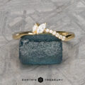 The "Liana" band in 18k yellow gold, featuring a marquise diamond and round accent stones, is shown on a rough blue stone with a geometric background and the text "EARTH'S TREASURY" below.