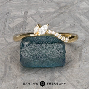 The "Liana" band in 18k yellow gold, featuring a marquise diamond and round accent stones, is shown on a rough blue stone with a geometric background and the text "EARTH'S TREASURY" below.