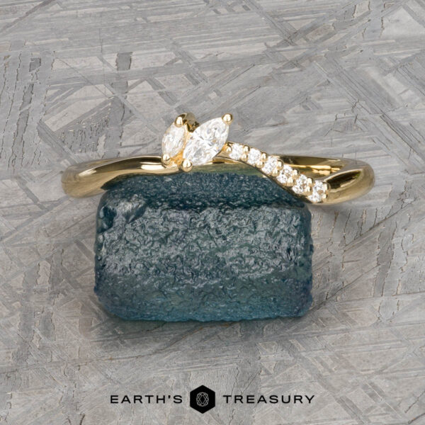 The "Liana" band in 18k yellow gold The "Liana" band in 18k yellow gold, featuring a marquise diamond and round accent stones, is shown on a rough blue stone with a geometric background and the text "EARTH'S TREASURY" below.