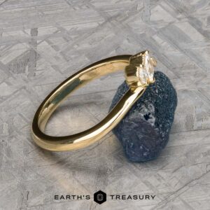The "Liana" band in 18k yellow gold, featuring a clear gemstone, is displayed against a rugged dark mineral on a textured gray surface, with the Earth's Treasury logo below.