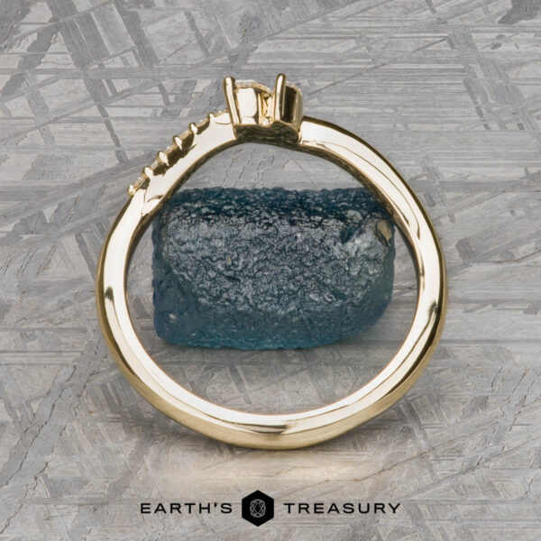 The "Liana" band in 18k yellow gold The "Liana" band in 18k yellow gold stands upright, encircling a rough blue stone at the center, set against a textured metallic background with "EARTH'S TREASURY" beneath.