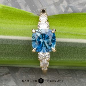 The "Celosia" ring in 14k yellow gold, featuring a 1.86-carat Montana sapphire center and accented by smaller clear stones, is displayed on a green leaf against a metallic backdrop.