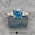 The "Celosia" ring in 14k yellow gold, featuring a 1.86-carat square-cut Montana sapphire center and round side stones, is displayed on a matching blue textured surface with "Earth's Treasury" at the bottom.
