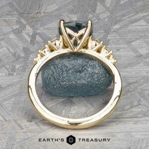 Gold ring named "Celosia" in 14k yellow gold with a 1.86-carat Montana sapphire, displayed on a textured surface background. The name "Earth's Treasury" appears at the bottom.