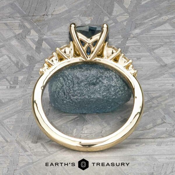 The "Celosia" ring in 14k yellow gold with 1.86-carat Montana sapphire Gold ring named "Celosia" in 14k yellow gold with a 1.86-carat Montana sapphire, displayed on a textured surface background. The name "Earth's Treasury" appears at the bottom.