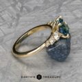 The "Celosia" ring, crafted in 14k yellow gold, displays a 1.86-carat Montana sapphire at the center, surrounded by multiple smaller clear stones on the band and rests on a metallic surface.