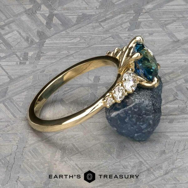 The "Celosia" ring in 14k yellow gold with 1.86-carat Montana sapphire The "Celosia" ring, crafted in 14k yellow gold, displays a 1.86-carat Montana sapphire at the center, surrounded by multiple smaller clear stones on the band and rests on a metallic surface.
