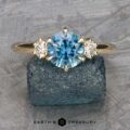 The "Clary" ring in 14k yellow gold features a 1.43-carat round Montana sapphire center stone and two smaller clear side stones, displayed on rough blue mineral with the Earth's Treasury logo beneath.