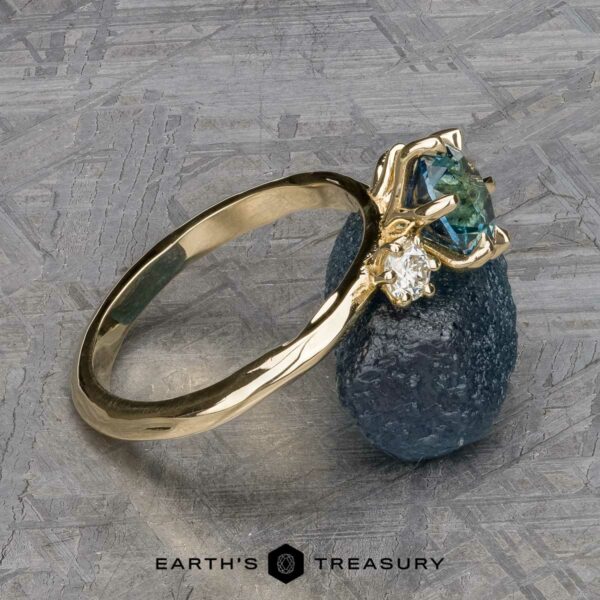 The "Clary" ring in 14k yellow gold with 1.43-Carat Montana Sapphire The "Clary" ring in 14k yellow gold, featuring a 1.43-carat Montana sapphire and a small clear stone, is shown resting on a rough blue mineral with the Earth's Treasury logo at the bottom.