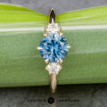 The "Clary" ring features a 1.43-carat oval Montana sapphire set in 14k yellow gold, accented by two round white diamonds, and is elegantly displayed on a green leaf background.