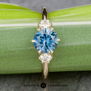 The "Clary" ring features a 1.43-carat oval Montana sapphire set in 14k yellow gold, accented by two round white diamonds, and is elegantly displayed on a green leaf background.