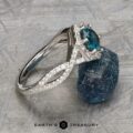 The "Graciella" ring in 14k white gold, featuring a 1.27-carat Montana sapphire and diamond accents, is shown beside a rough blue stone on a textured gray surface. Text at the bottom reads "Earth's Treasury.