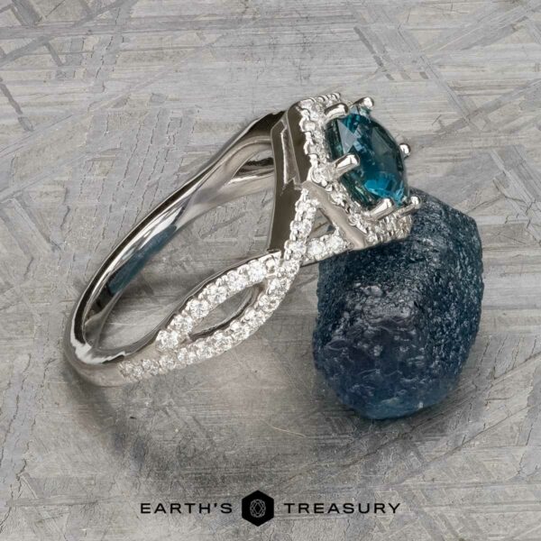 The "Graciella" ring in 14k white gold with 1.27-carat Montana sapphire The "Graciella" ring in 14k white gold, featuring a 1.27-carat Montana sapphire and diamond accents, is shown beside a rough blue stone on a textured gray surface. Text at the bottom reads "Earth's Treasury.