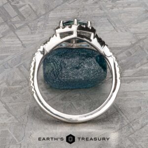 The "Graciella" ring in 14k white gold with a 1.27-carat Montana sapphire stands upright on a textured gray surface, with the "EARTH'S TREASURY" logo visible at the bottom.