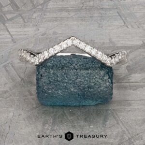 The "Graciella" band in 14k white gold, featuring a chevron-shaped design, is displayed upright on a rough blue gemstone against a textured gray background with the "Earth's Treasury" logo below.