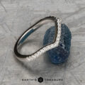 The "Graciella" band in 14k white gold, featuring a curved, textured design, is displayed leaning against a rough blue gemstone on a patterned surface. The logo "Earth's Treasury" appears at the bottom.