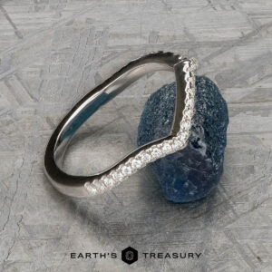 The "Graciella" band in 14k white gold, featuring a curved, textured design, is displayed leaning against a rough blue gemstone on a patterned surface. The logo "Earth's Treasury" appears at the bottom.